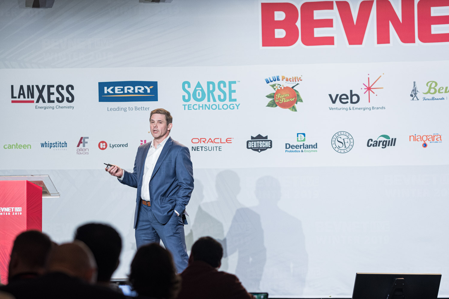 A man in a navy suit stands on stage in front of a LANXESS, Kerry and Sorse sponsor wall with the audience in silhouette.