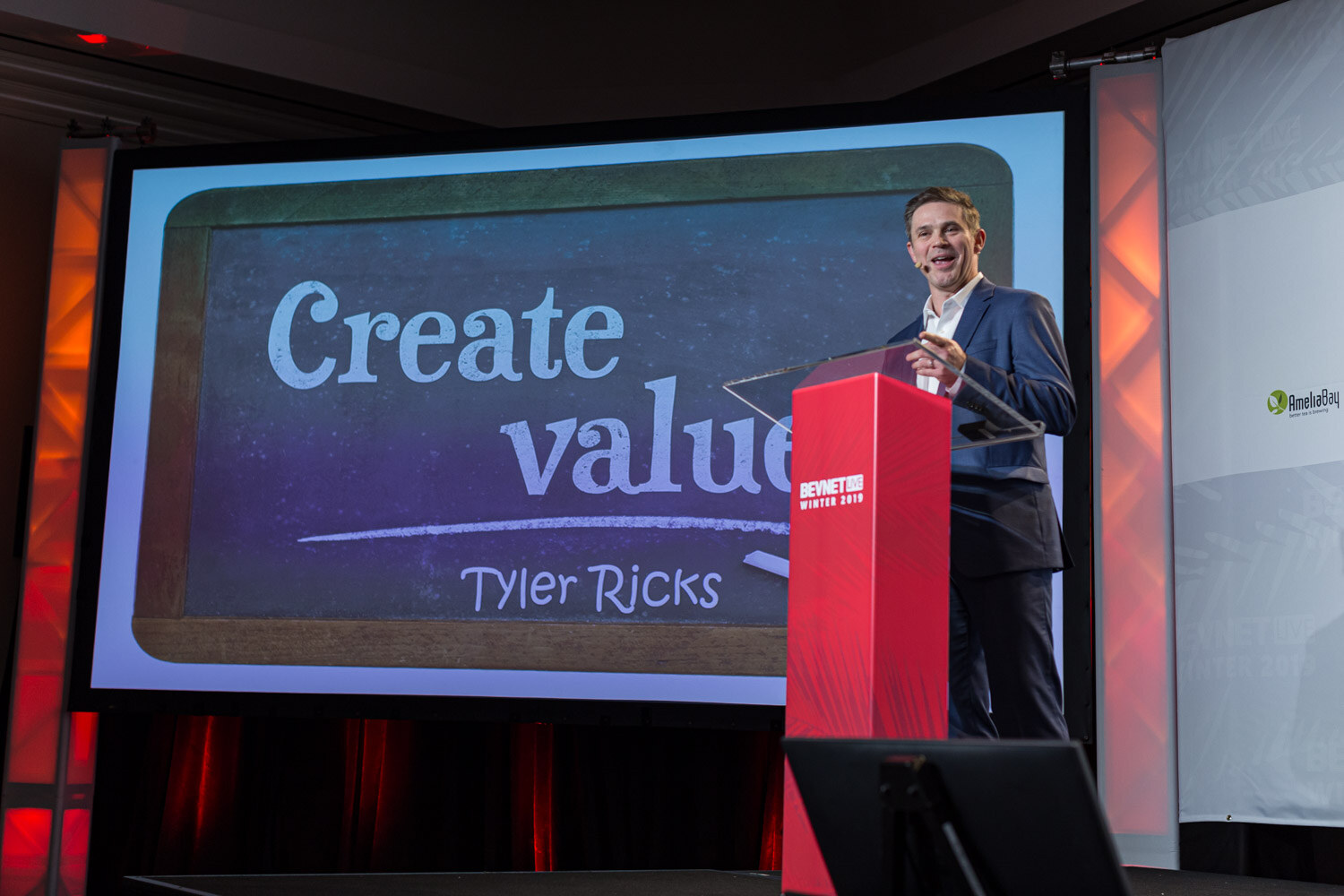 A man in a navy suit stands by a red lectern beside a chalkboard-style slide reading Create value, Tyler Ricks.