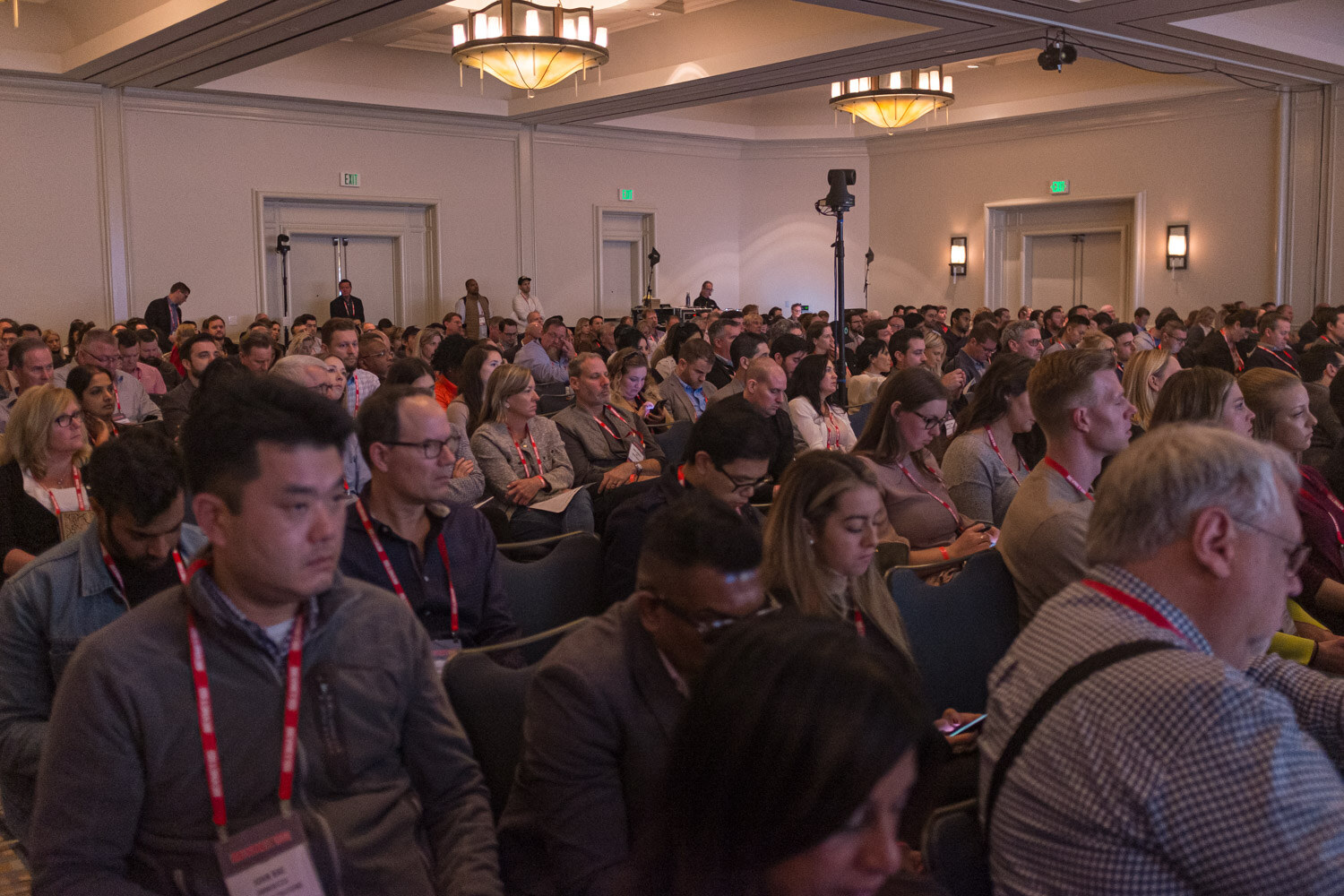 Rows of attendees with red lanyards sit facing the stage in a chandeliered ballroom.