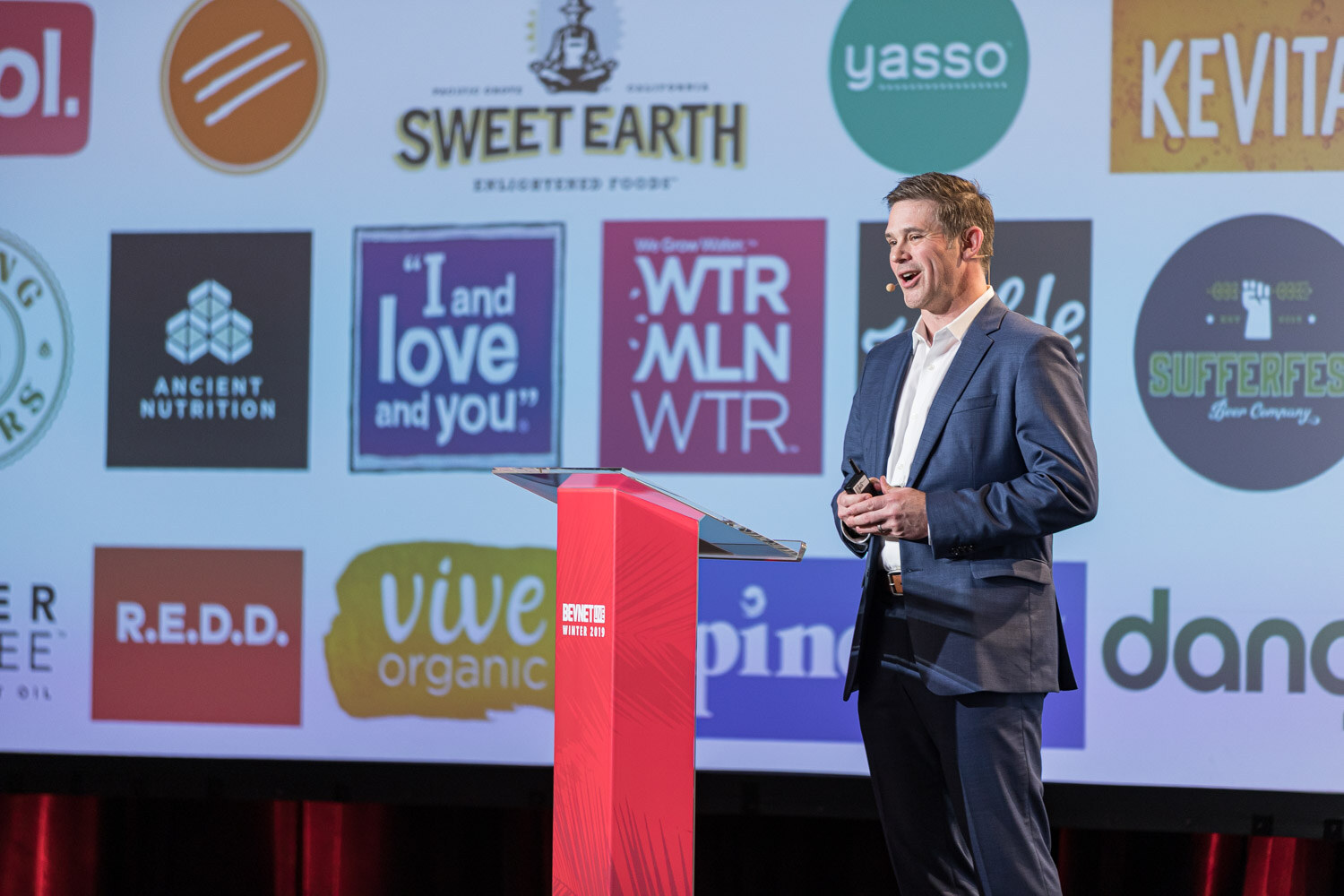 A man in a navy suit stands at a lectern in front of a large screen showing logos including Sweet Earth, Kevita, R.E.D.D., Vive Organic and Ancient Nutrition.