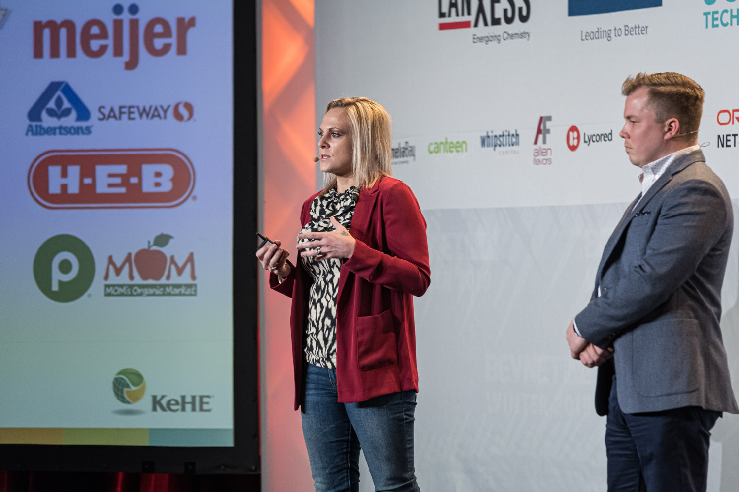 A woman in a red jacket and a man in a grey blazer stand on stage beside a screen showing Meijer, Albertsons, Safeway, H-E-B and KeHE logos.
