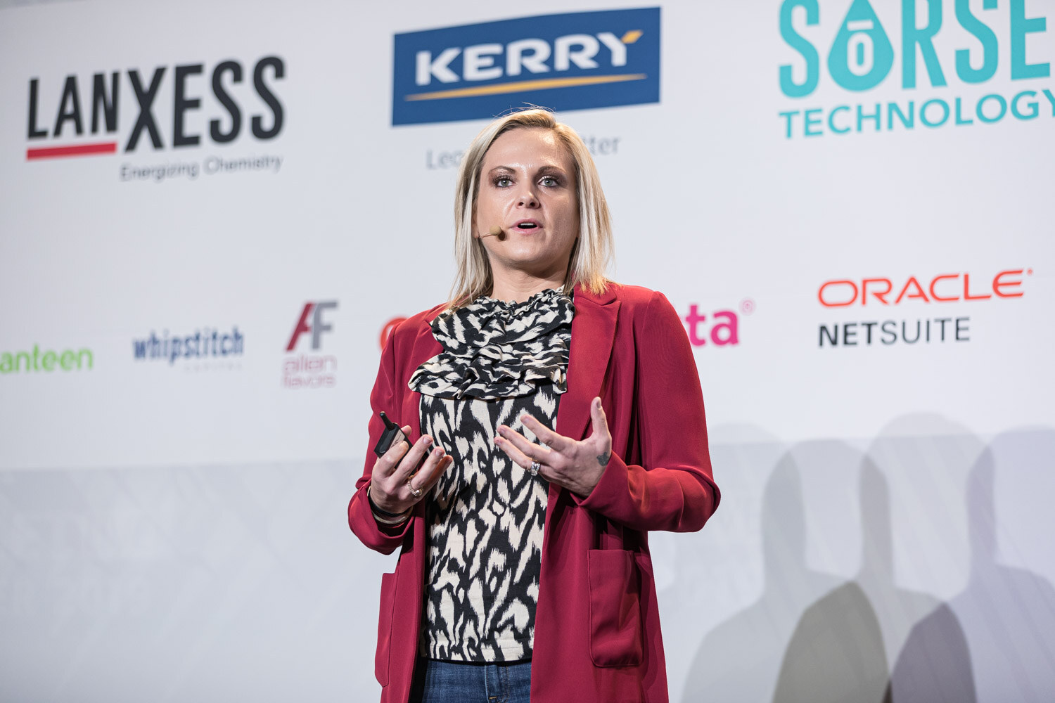 A woman in a red jacket with a black and white top speaks into a headset microphone.
