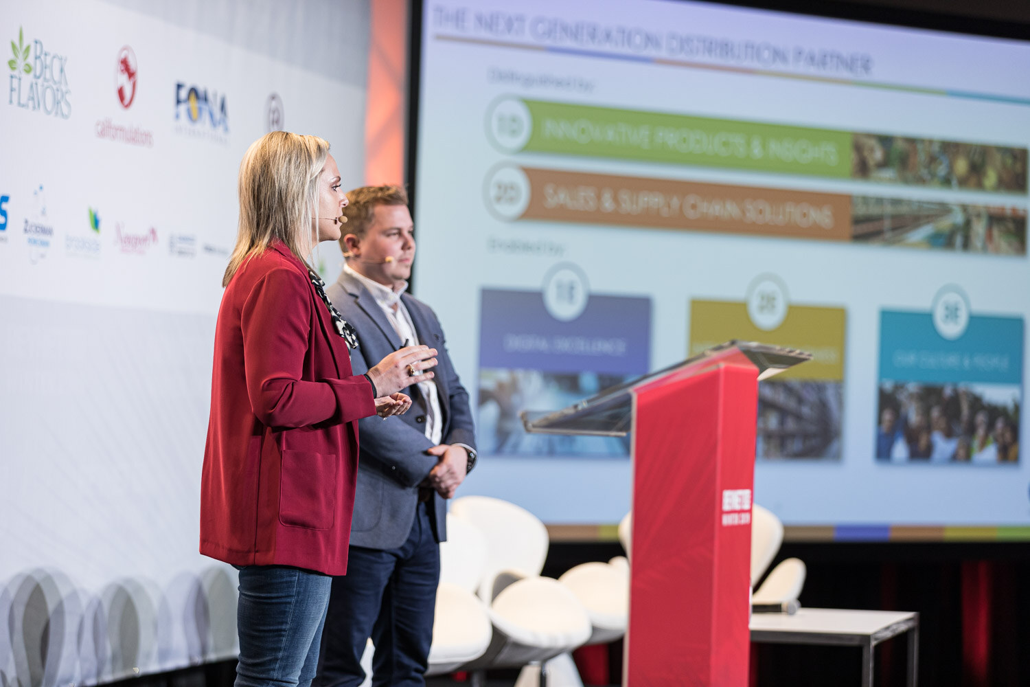 A woman in a red jacket and a man in a grey blazer stand beside a lectern and a screen titled The Next Generation Distribution Partner.