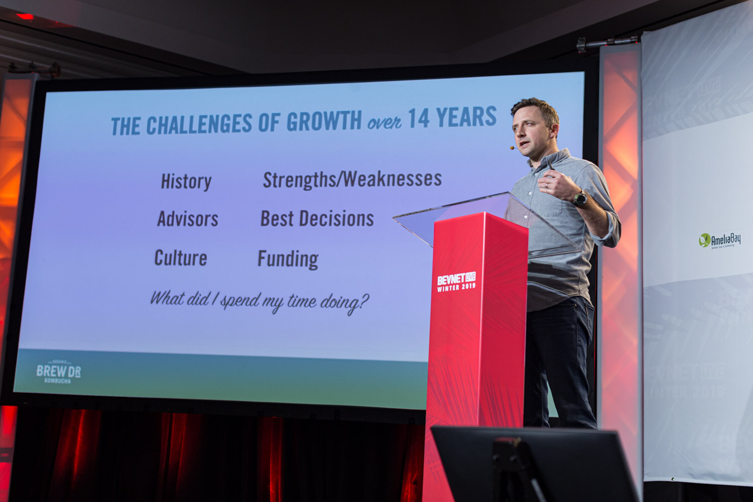 A man in a grey shirt stands at a red lectern beside a screen listing History, Advisors, Culture, Strengths/Weaknesses, Best Decisions and Funding.