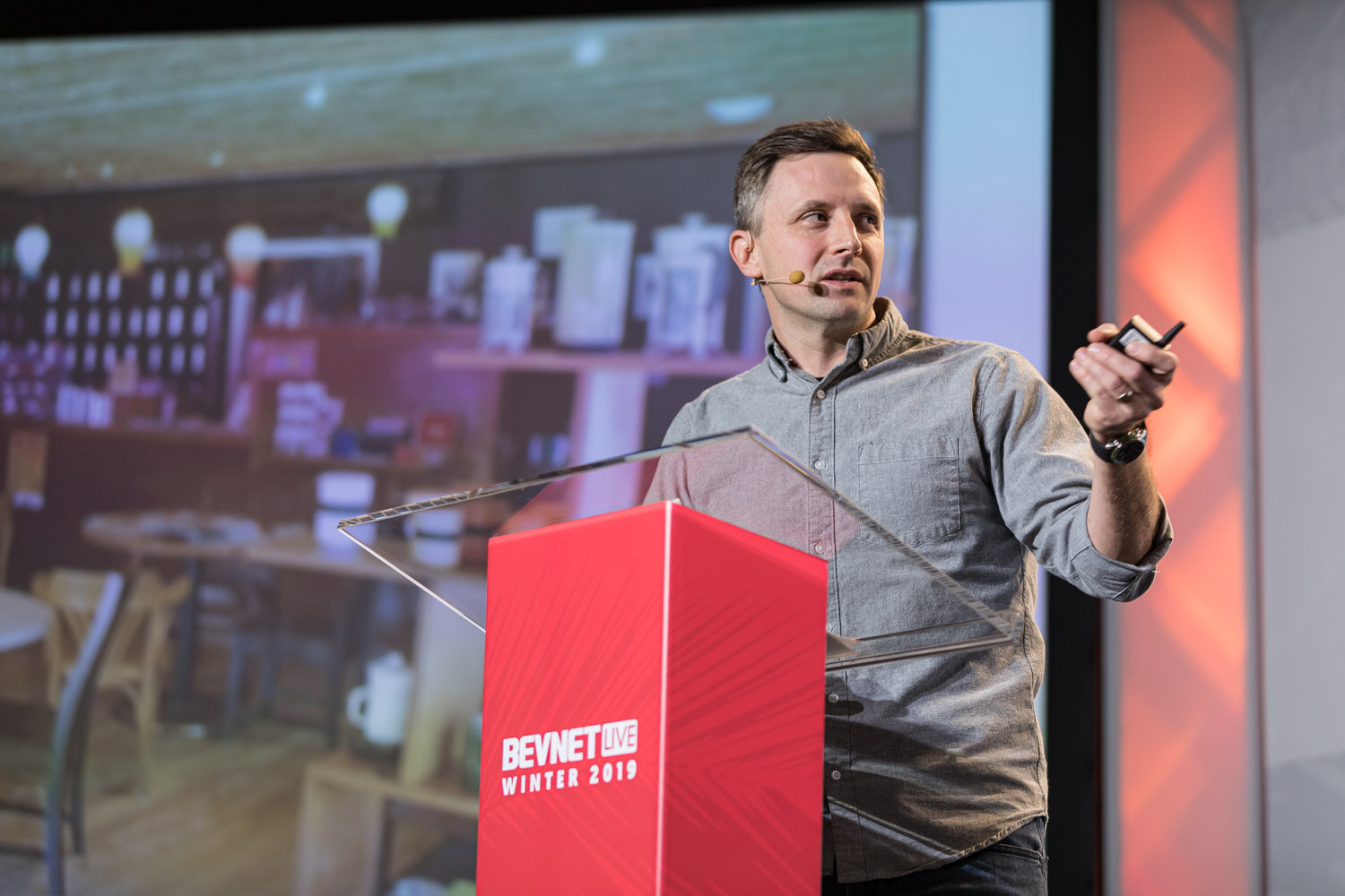 A man in a grey shirt holds a clicker at a red lectern with a photo of shelves and tables projected behind him.