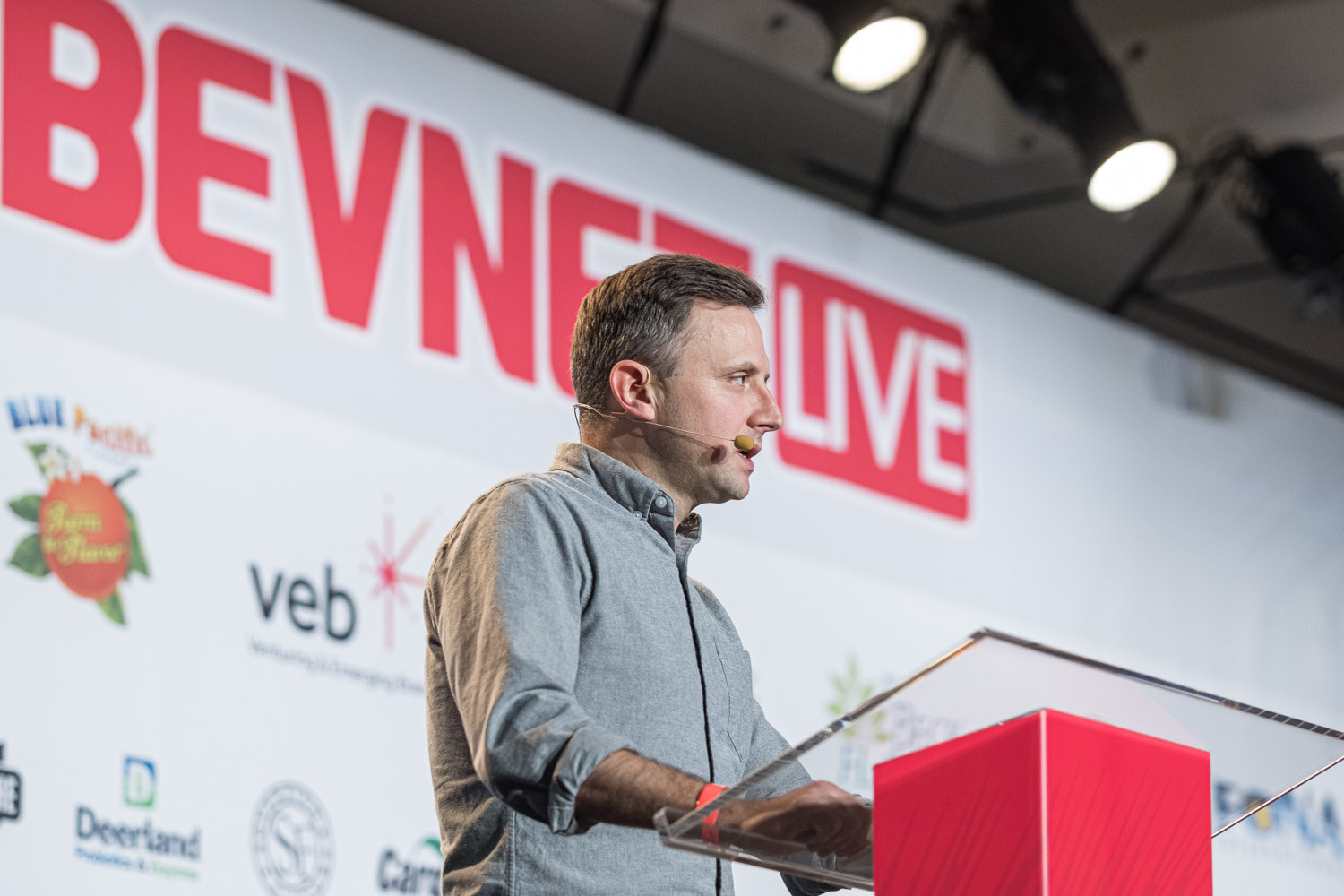 A man in a grey shirt stands at a lectern with the BevNET Live backdrop and a sponsor wall behind him.
