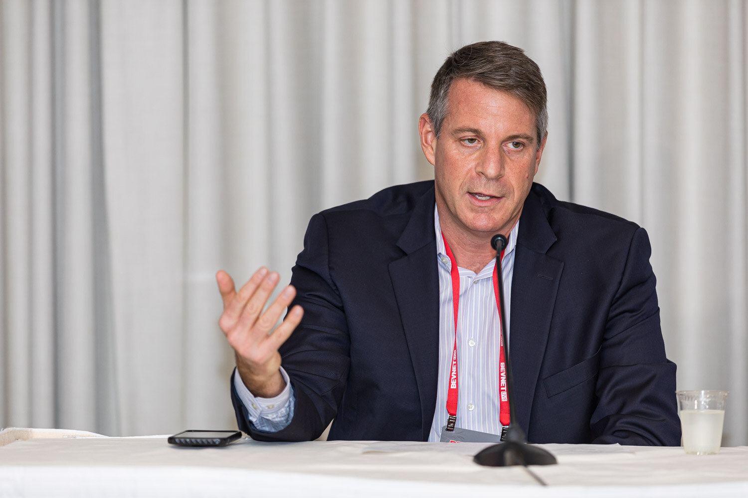 A man in a navy blazer with a lanyard speaks at a table with microphones in front of a curtain.