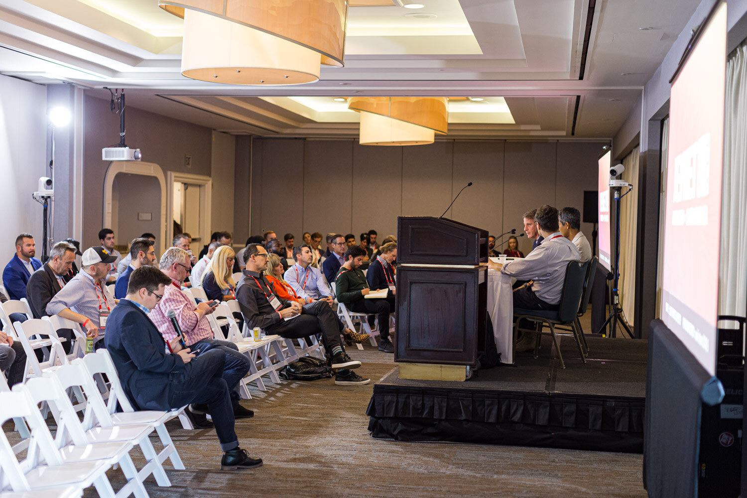 Rows of white folding chairs face a podium and stage in a hotel meeting room with an attentive audience.