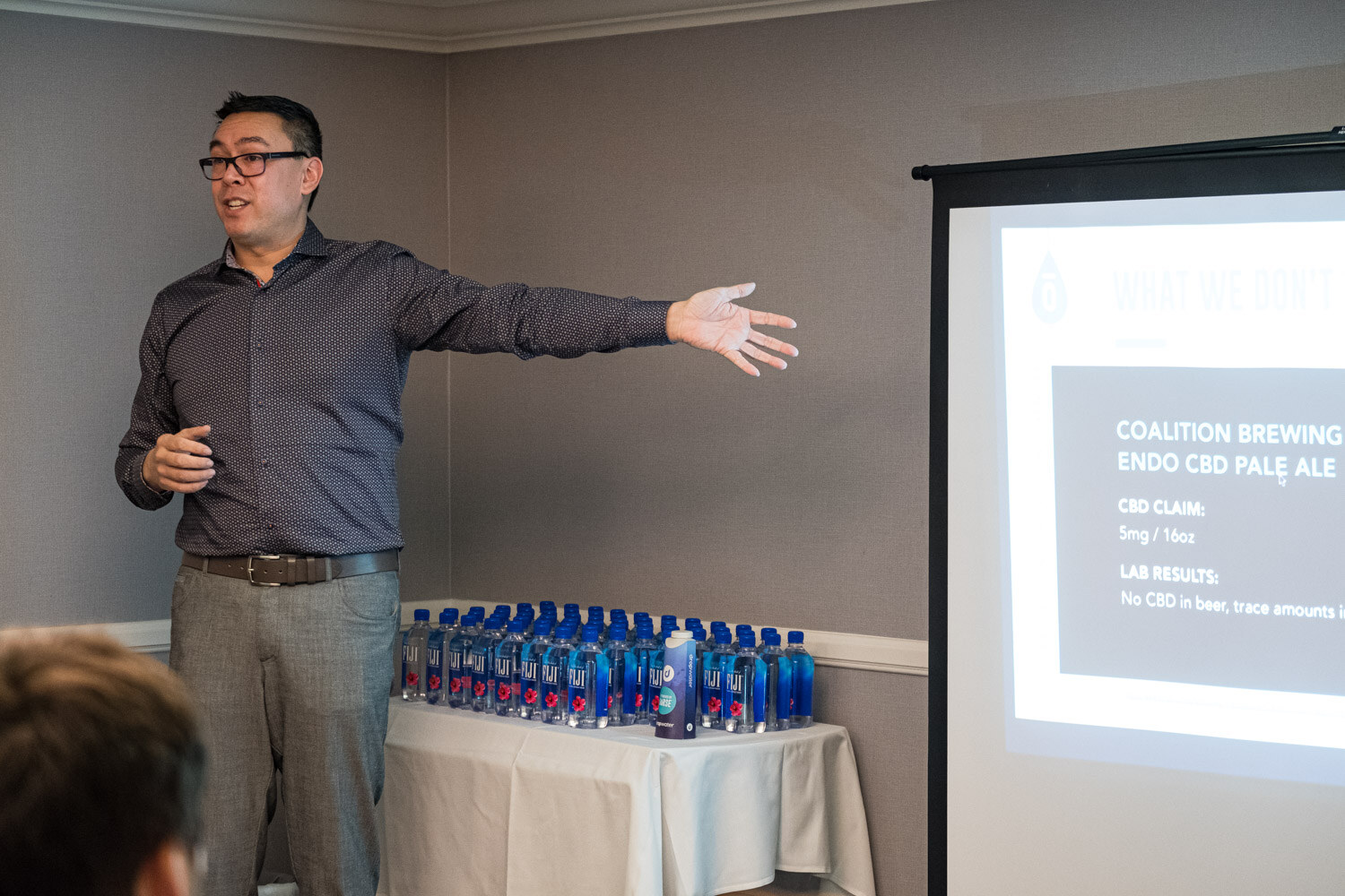 A man in glasses and a grey shirt points toward a projection screen listing a CBD pale ale, beside a table of water bottles.