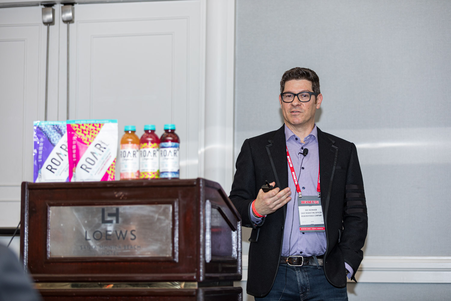A man in glasses and a dark blazer stands beside a Loews podium topped with ROAR boxes and bottles.