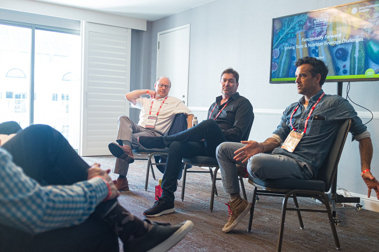 Three men with lanyards sit on folding chairs in a hotel meeting room with a screen reading Functionally Forward in the corner.