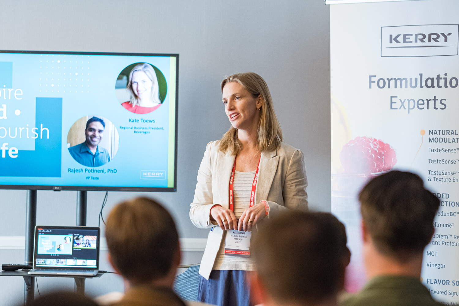 A bearded man with a red lanyard gestures in front of a Kerry Formulation Experts banner while a woman in a beige blazer stands beside him.