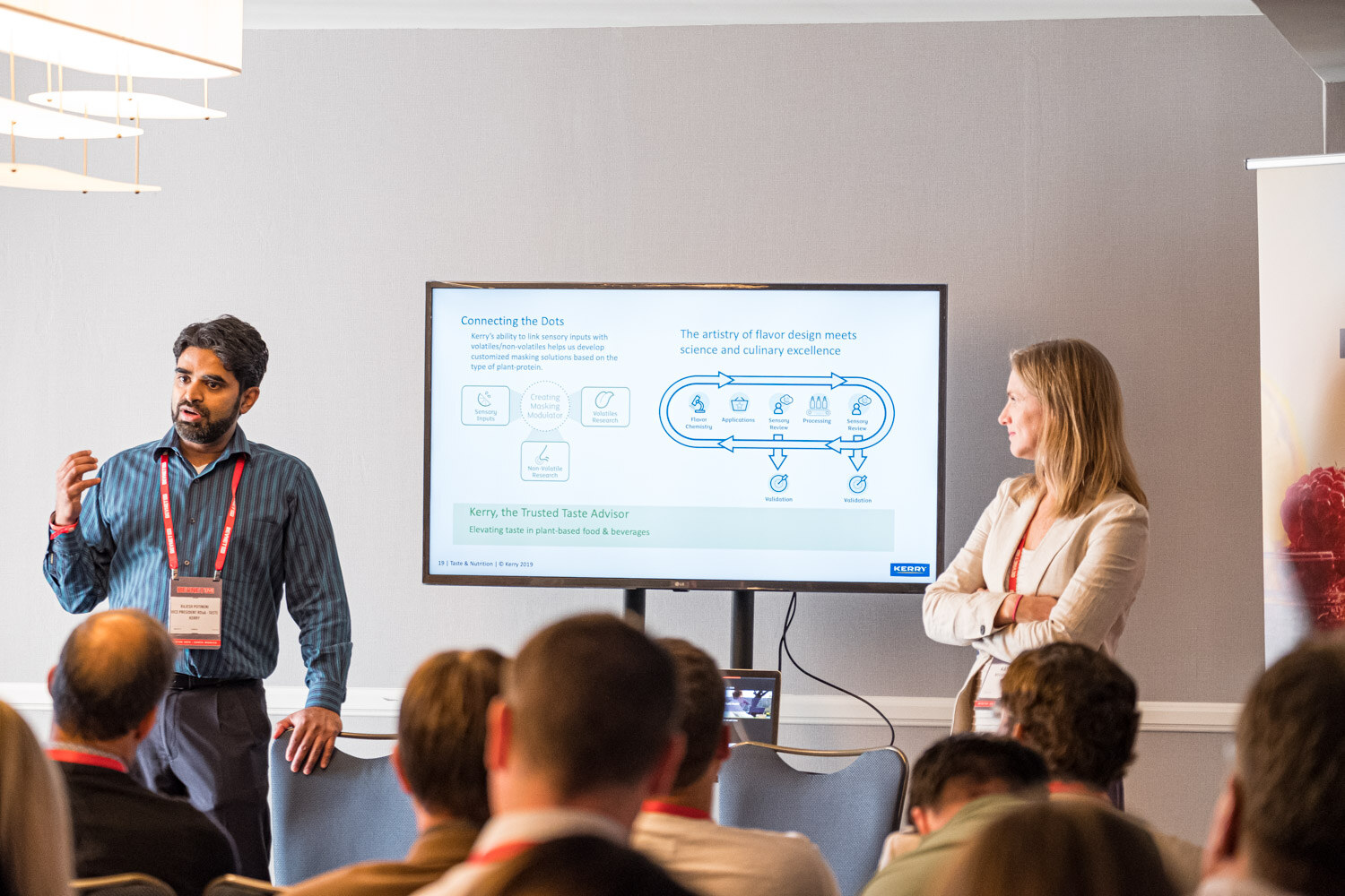 A man in a striped shirt and a woman in a blazer present beside a screen showing a flavor and taste diagram while attendees watch.
