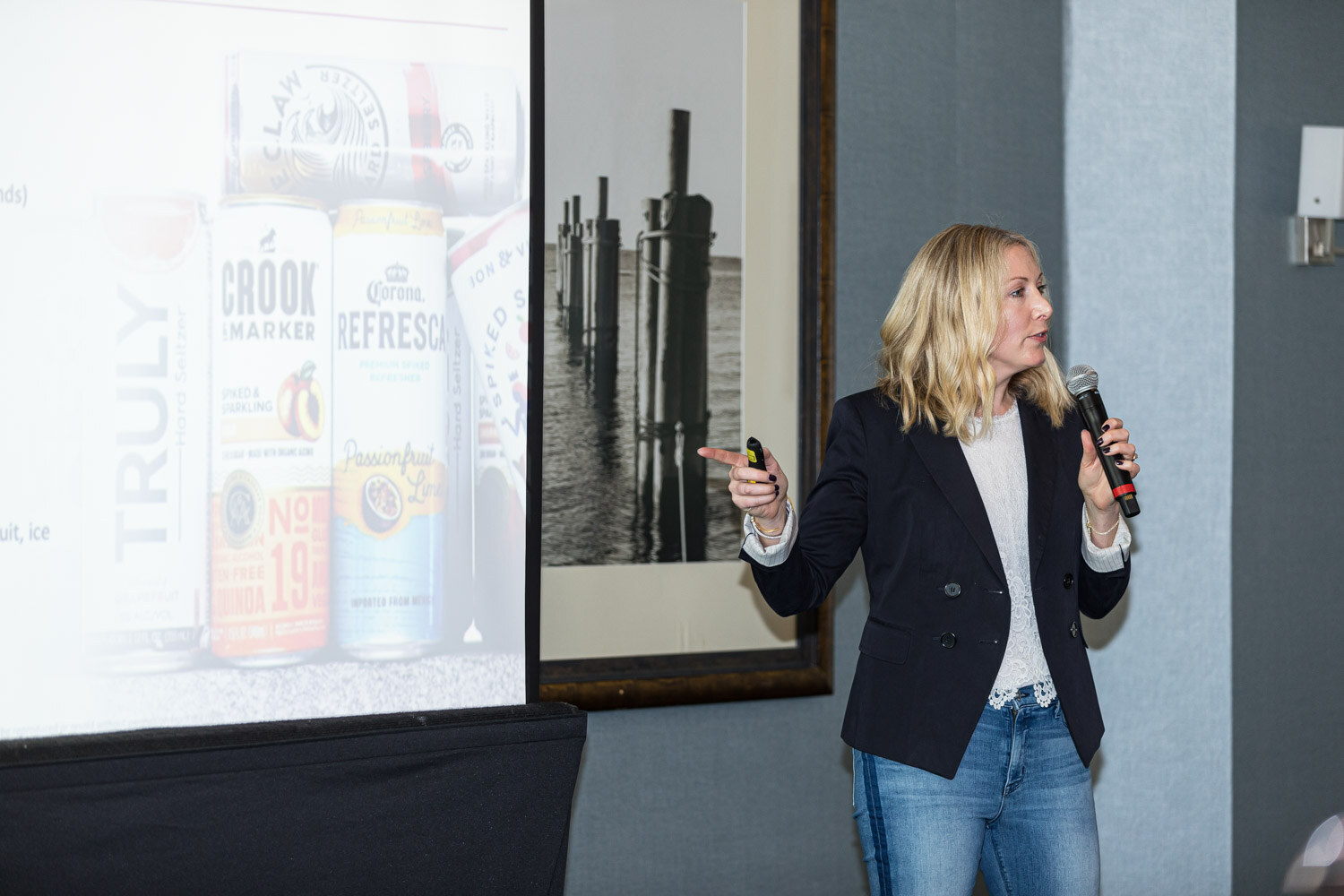 A woman in a navy blazer holds a microphone and a clicker next to a screen showing TRULY and Crook and Marker cans.