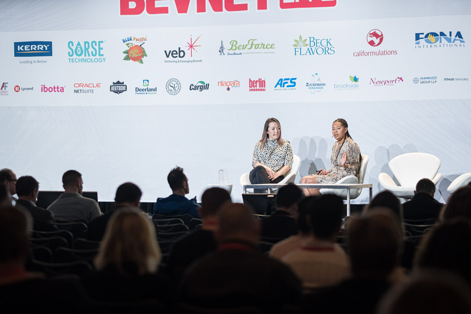 A wide shot of the stage with two women seated on white chairs in front of a sponsor wall and silhouetted audience.