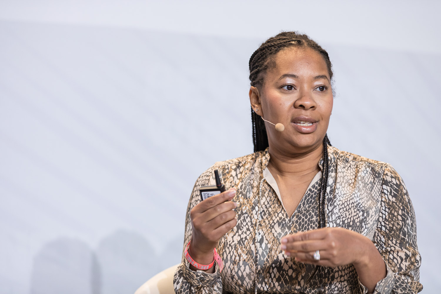 A woman in a snake-print blouse and a headset microphone speaks while holding a small card.