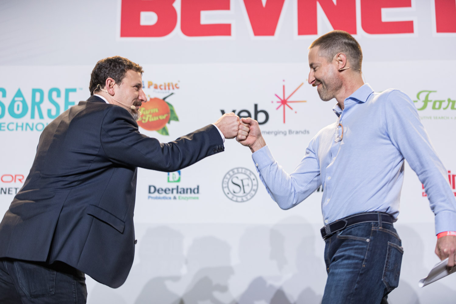 A man in a dark blazer and a man in a light blue shirt bump fists in front of the sponsor wall.
