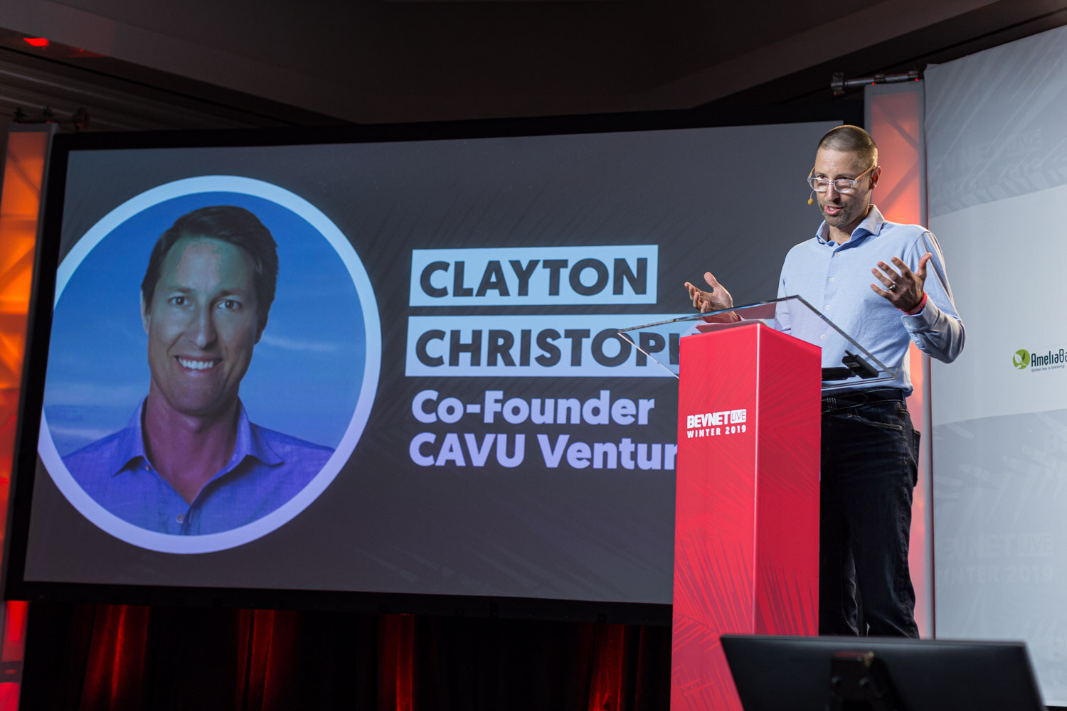 A man in glasses and a light blue shirt speaks at a red lectern beside a screen showing a portrait and the name Clayton Christopher, Co-Founder CAVU Venture.