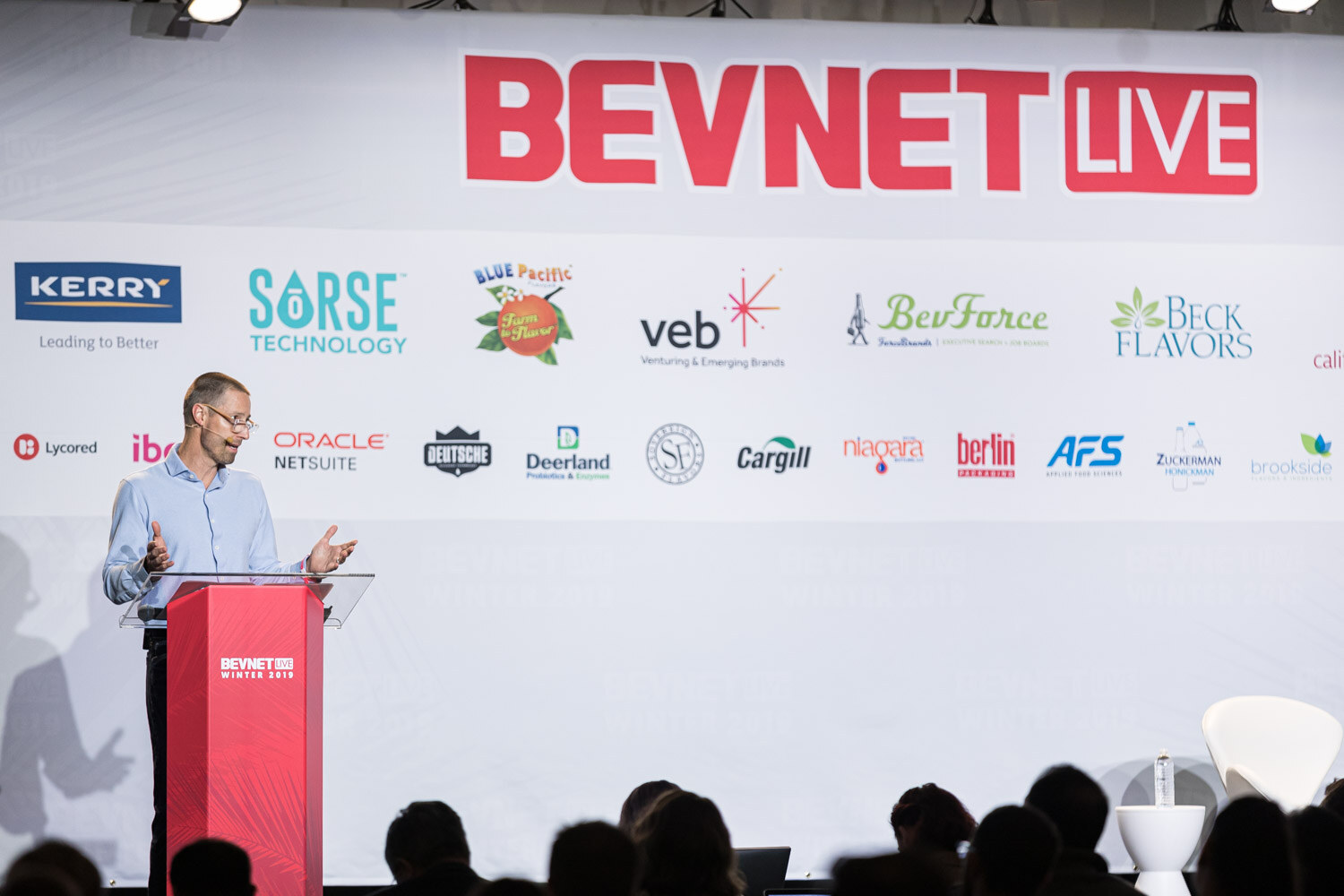 A man in a blue shirt at a red lectern speaks in front of the BevNET Live sponsor wall with the audience in silhouette.