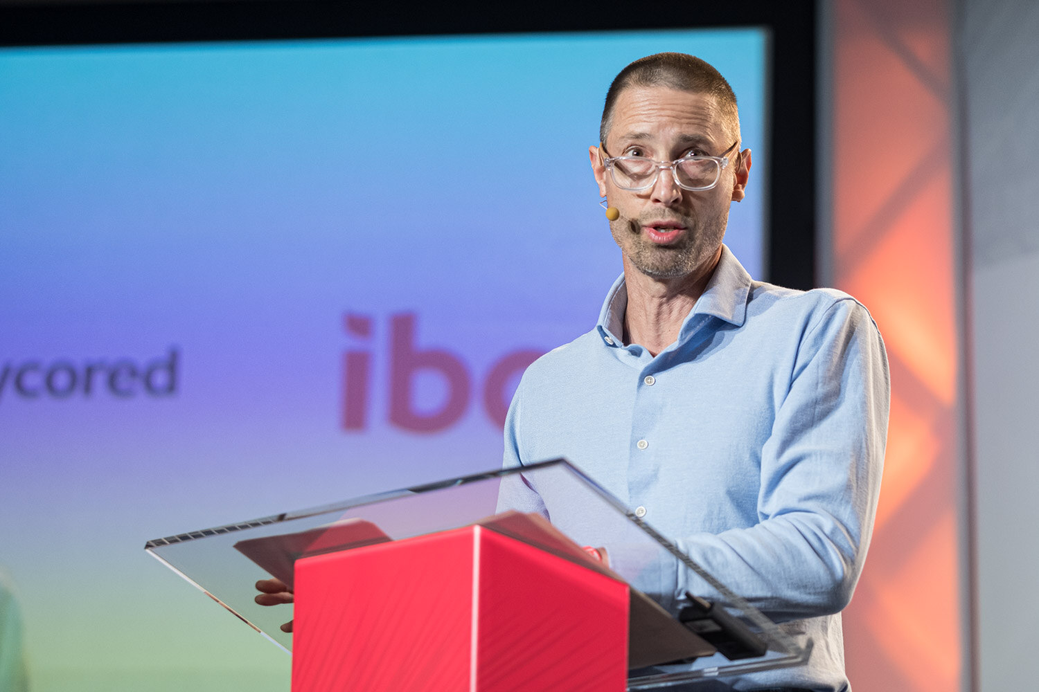 A man in glasses and a light blue shirt speaks into a headset microphone at a clear lectern in front of a colorful screen.