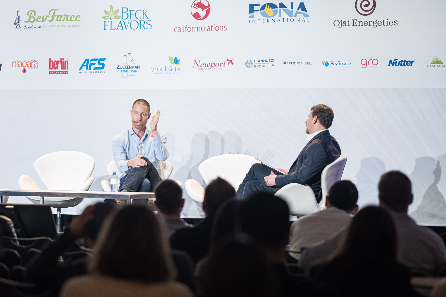 Silhouettes of the audience in the foreground face two seated speakers on a stage with a BevNET Live backdrop.