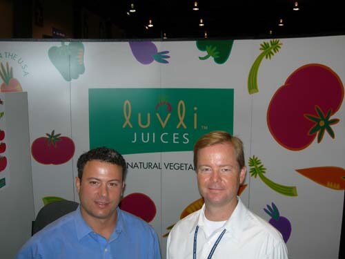 Two men stand side by side in front of a Luvli Juices sign with illustrated vegetables; one wears a blue button-up shirt and the other a white shirt with a lanyard.