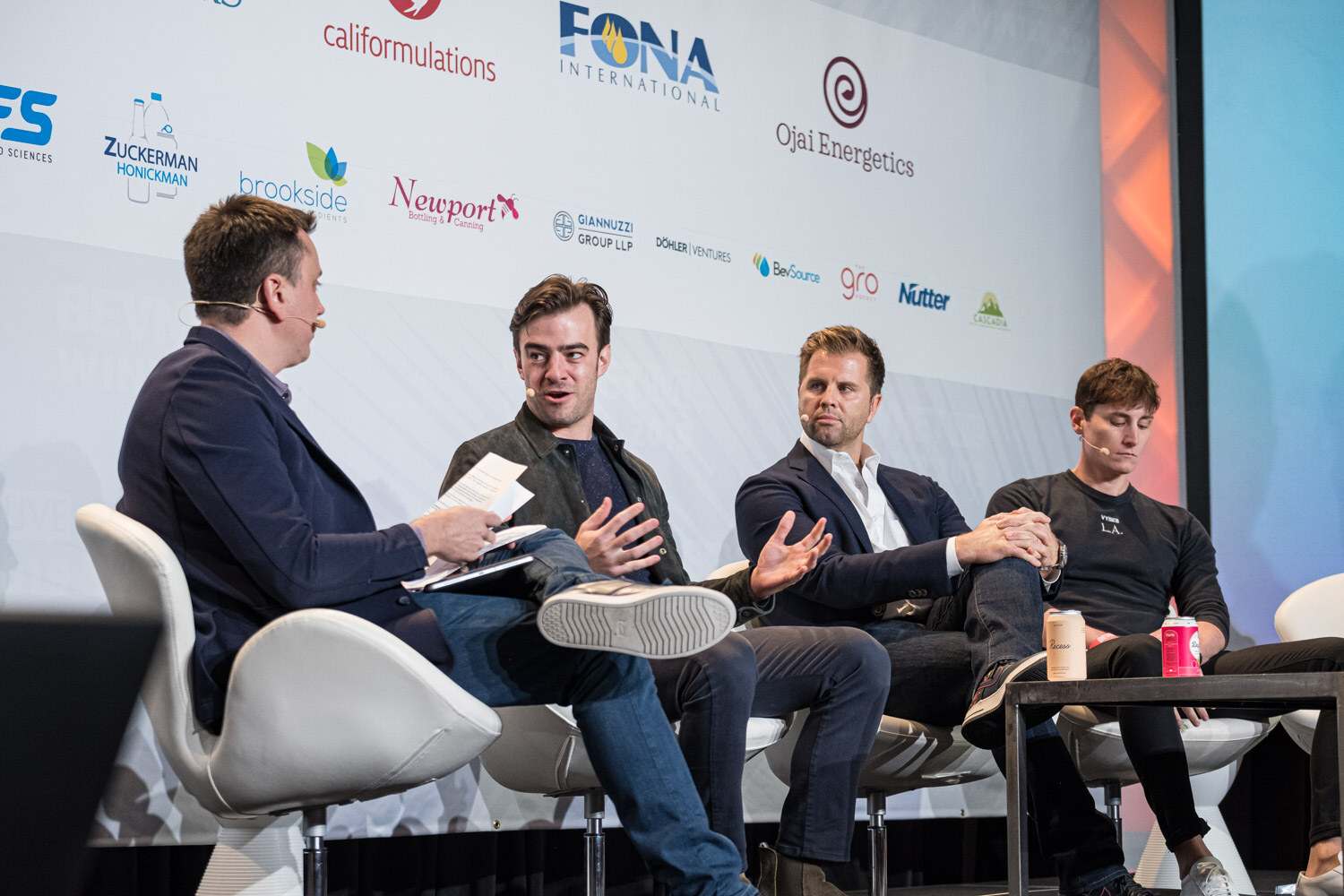 A man in a navy sweater holding notes sits beside three men with cans on the table in front of a sponsor wall.