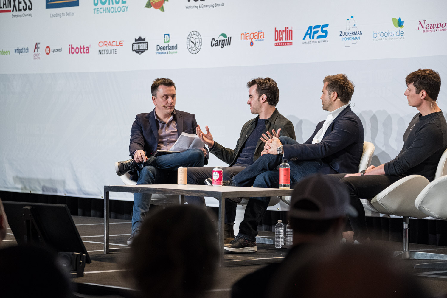 Four men on white chairs around a low table with cans on it in front of a sponsor wall.