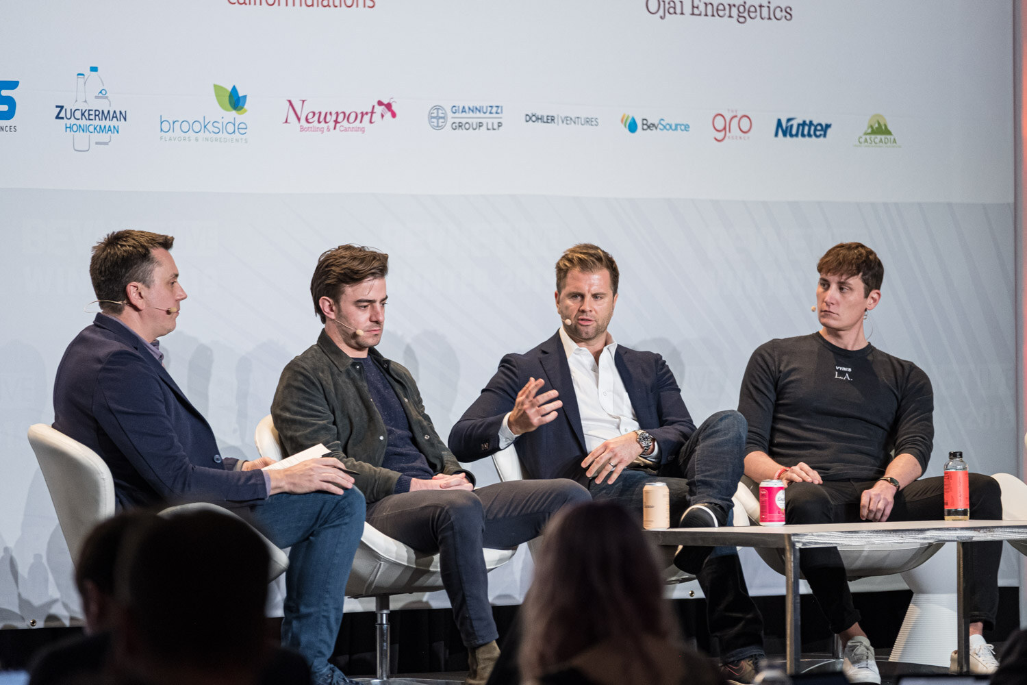 Four men seated on white chairs on stage, with cans and bottles on the table in front of them.