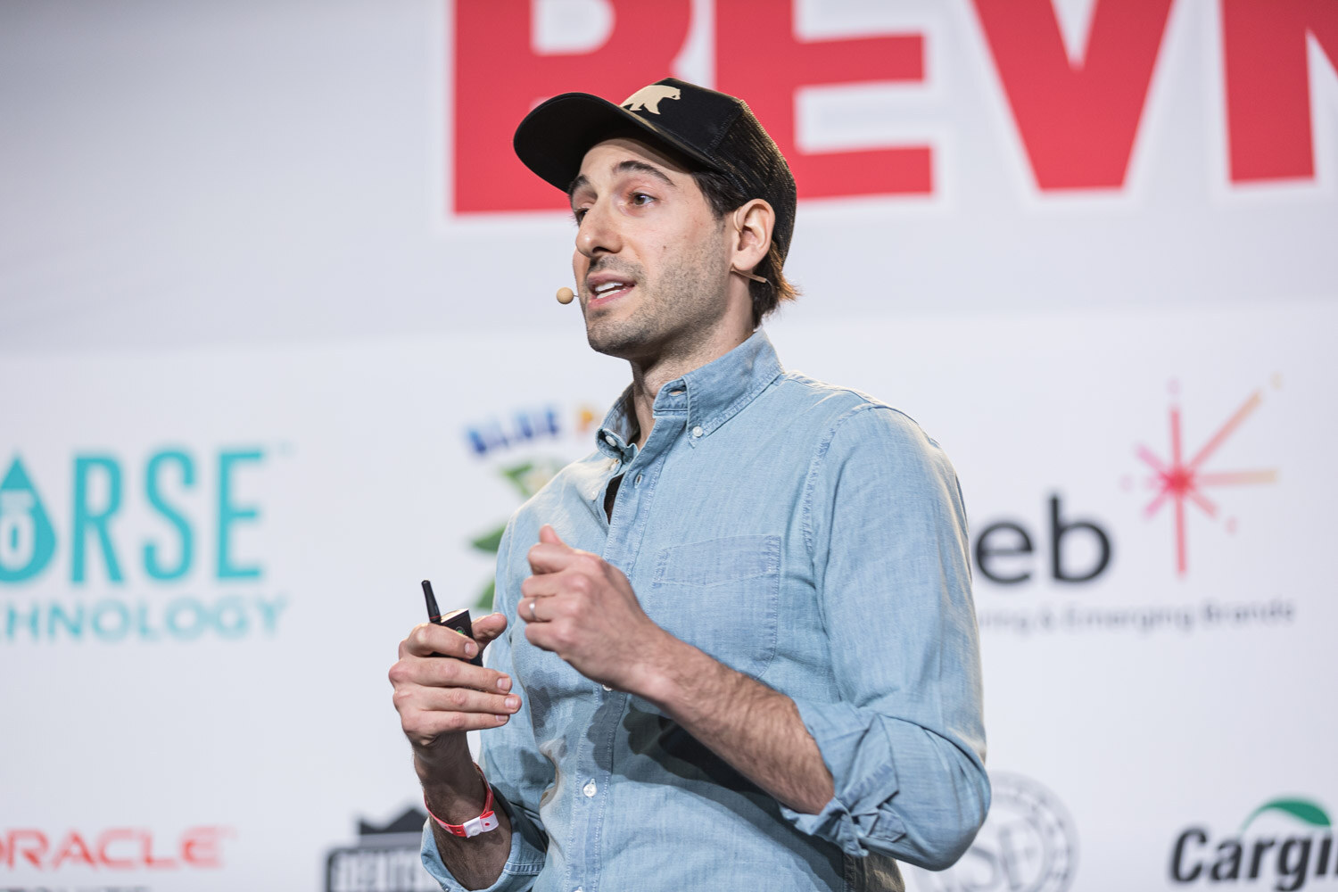 A man in a black cap and blue chambray shirt holding a clicker speaks in front of the sponsor wall.