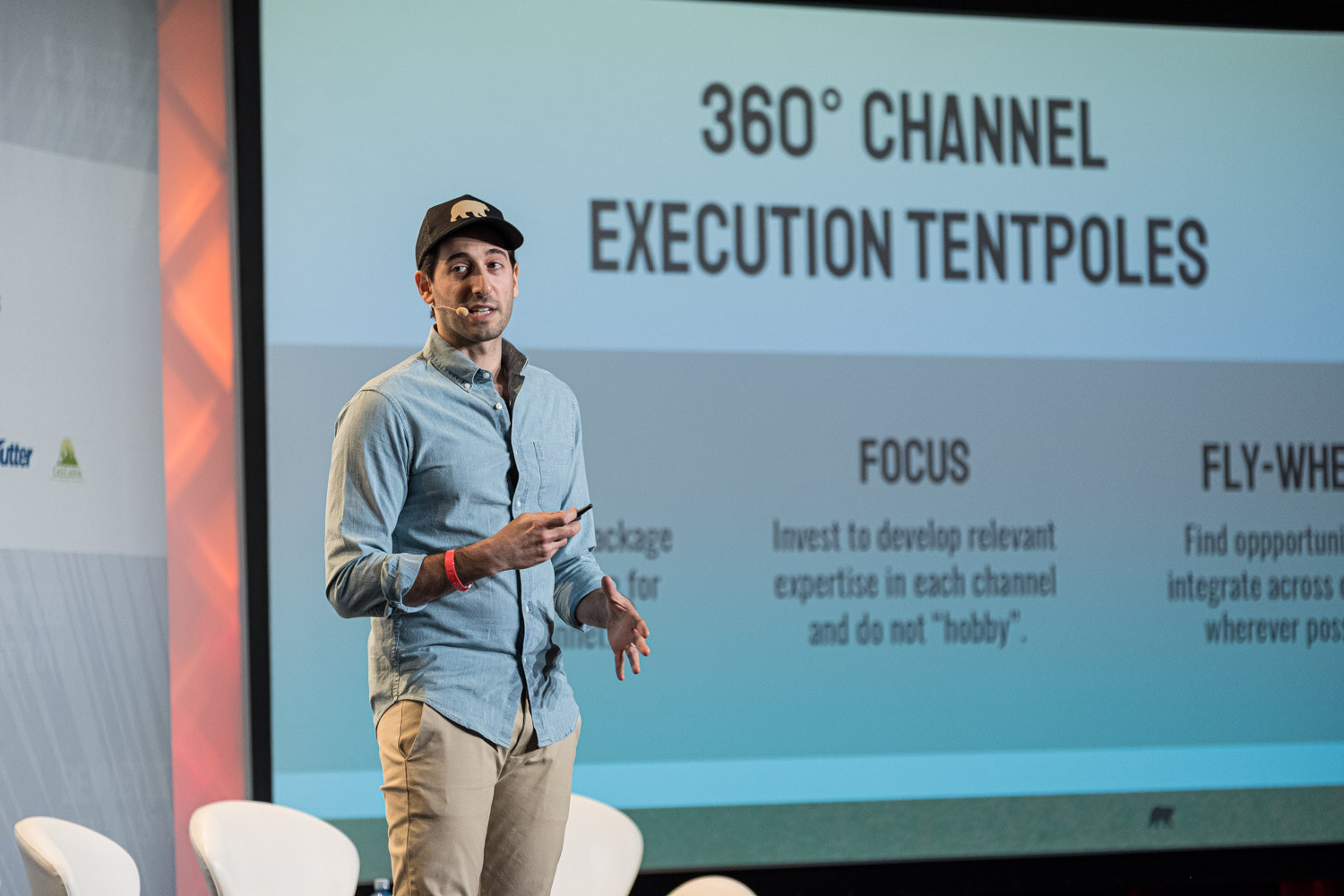 A man in a cap and chambray shirt stands before a screen reading 360 Channel Execution Tentpoles with Focus and Fly-Wheel sections.