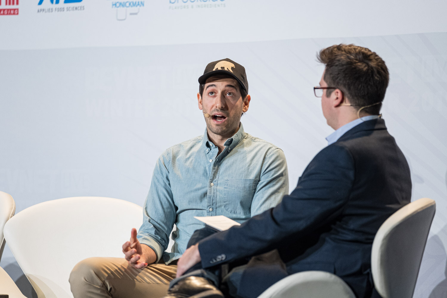 A man in a cap and light shirt talks with a man in a navy jacket and glasses while seated on white chairs.