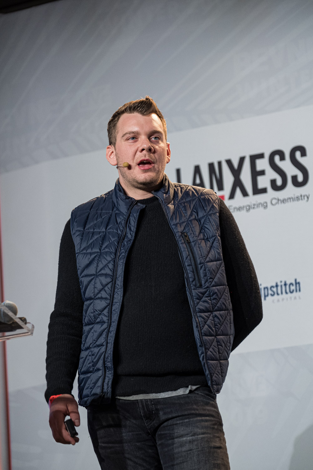 A man in a quilted navy vest and black sweater stands in front of a LANXESS and Kerry sponsor wall.