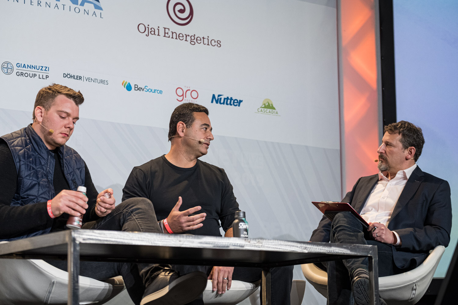 A man in a quilted vest and a man in a black shirt sit at a table beside a moderator holding a clipboard.