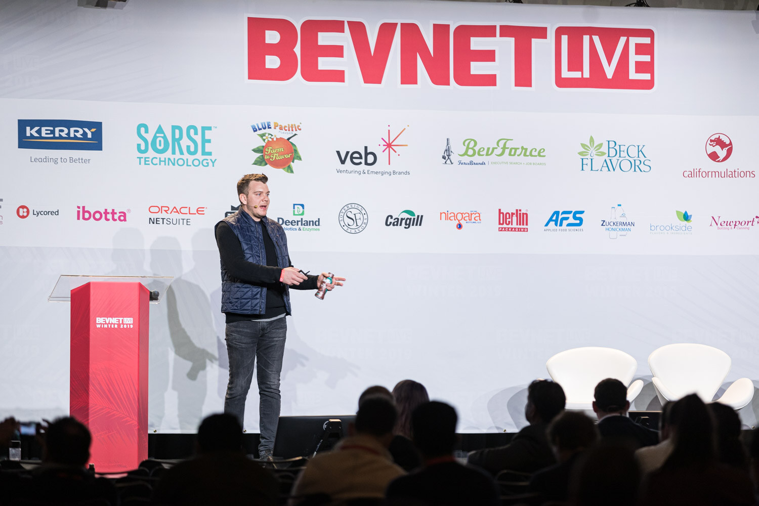 A wide view of a man in a vest presenting beside a red lectern with the audience in silhouette.