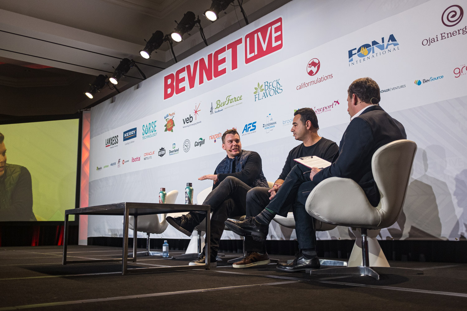 Three men sit on white chairs on a stage in front of a sponsor wall and a lit screen.