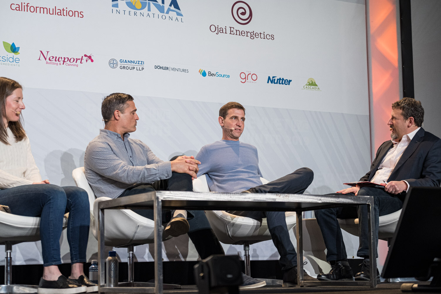 Four people sit on white chairs around a low table on stage in front of a sponsor wall.