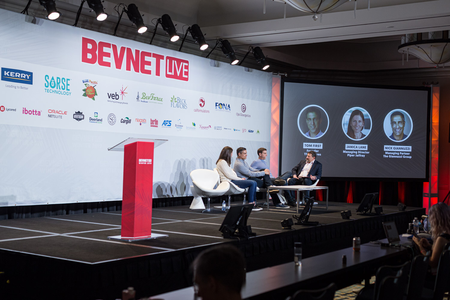 A wide shot of the stage with a red lectern, four panelists seated in white chairs and three portraits on the screen behind them.
