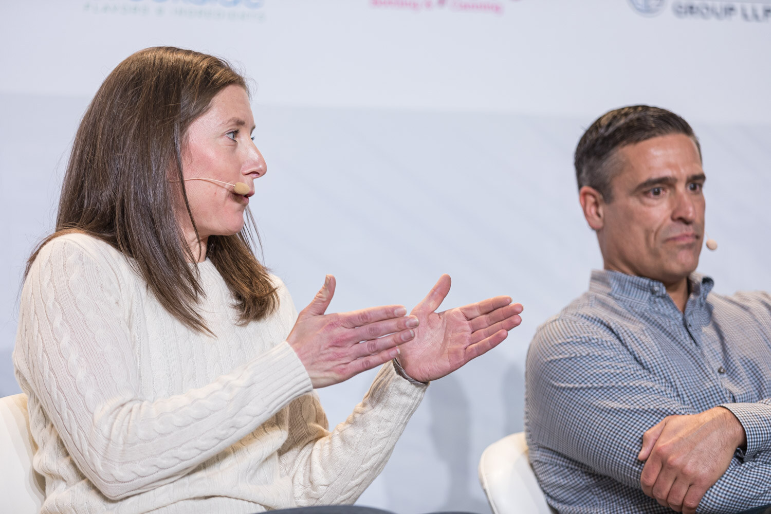 A woman in a cream sweater with her hands spread and a man in a checked shirt seated on white chairs on stage.