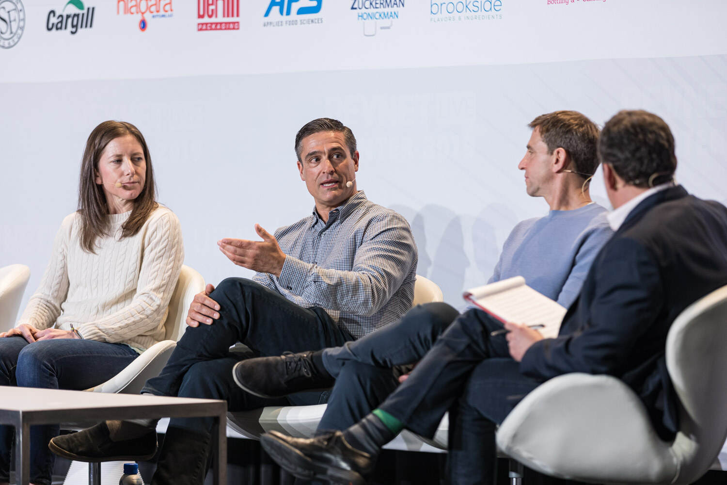 Four people on white chairs, one gesturing with an open hand, as a moderator holding papers watches.