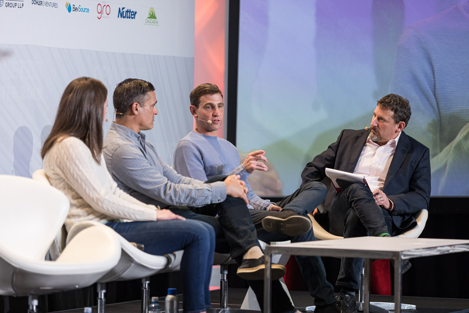 Four seated panelists, including a man in a dark jacket holding notes, on white chairs in front of the sponsor wall.