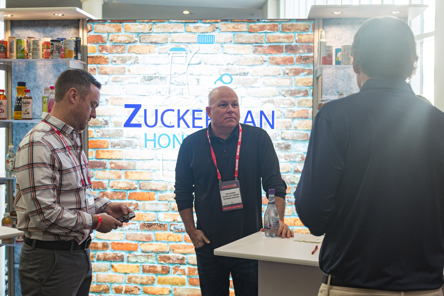 A man checks his phone and another stands beside a counter in front of a brick wall with a Zuckerman Honickman logo.
