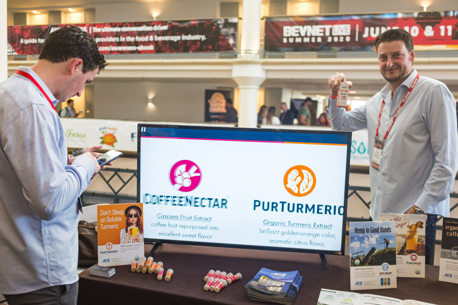 Two men with lanyards stand behind a table with a screen showing CoffeeNectar and PurTurmeric logos on the hotel atrium balcony.