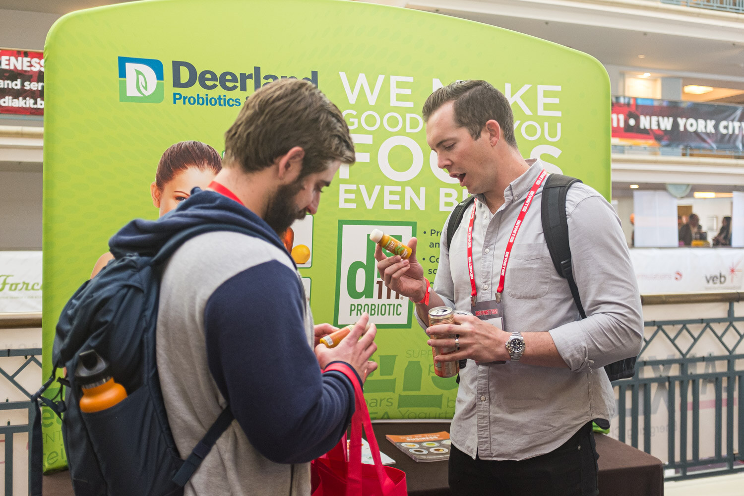 A man with a backpack bites into a sample while another attendee talks with him in front of a green Deerland Probiotics display.