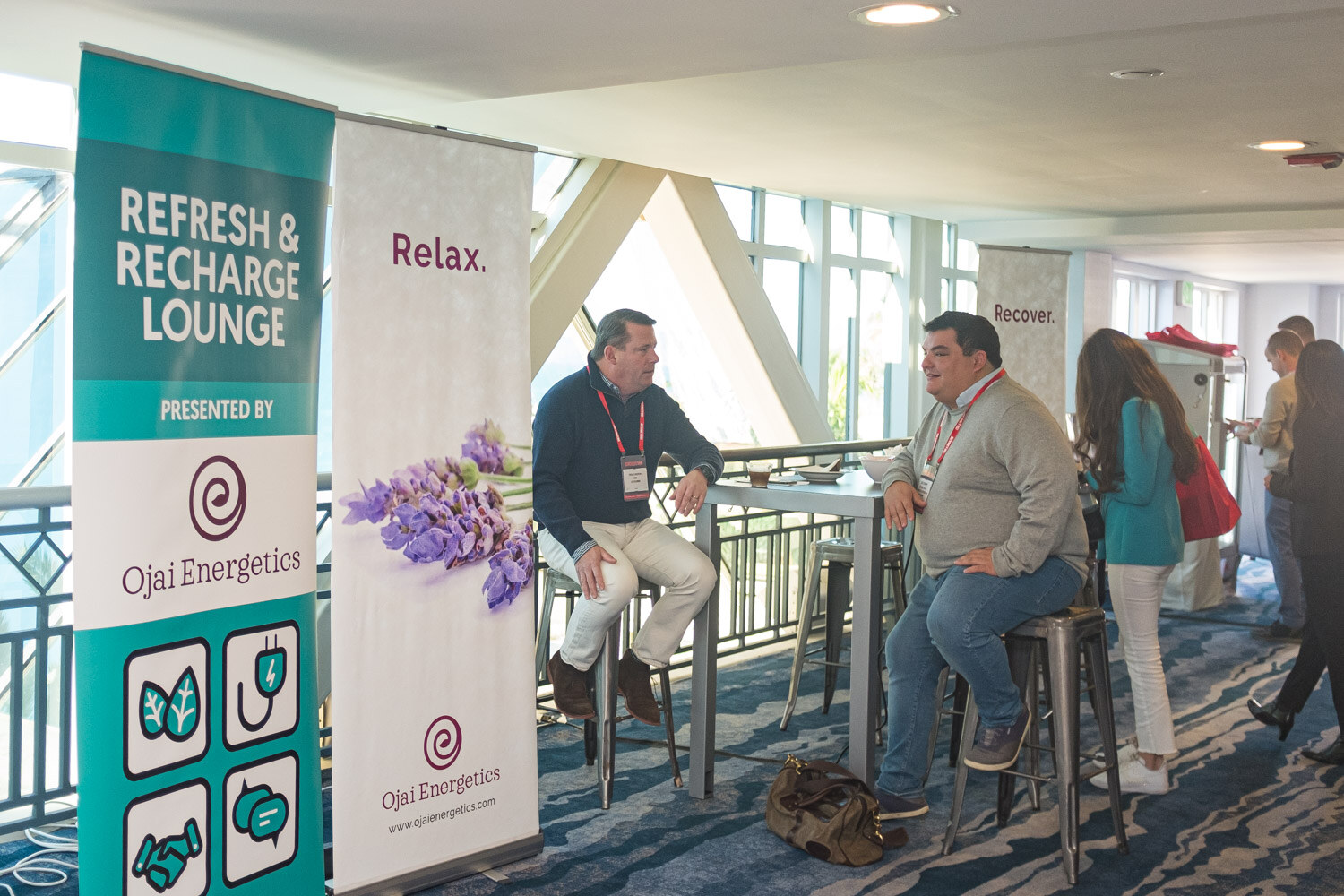 Two men sit at a high table beside Ojai Energetics banners reading Refresh and Recharge lounge.