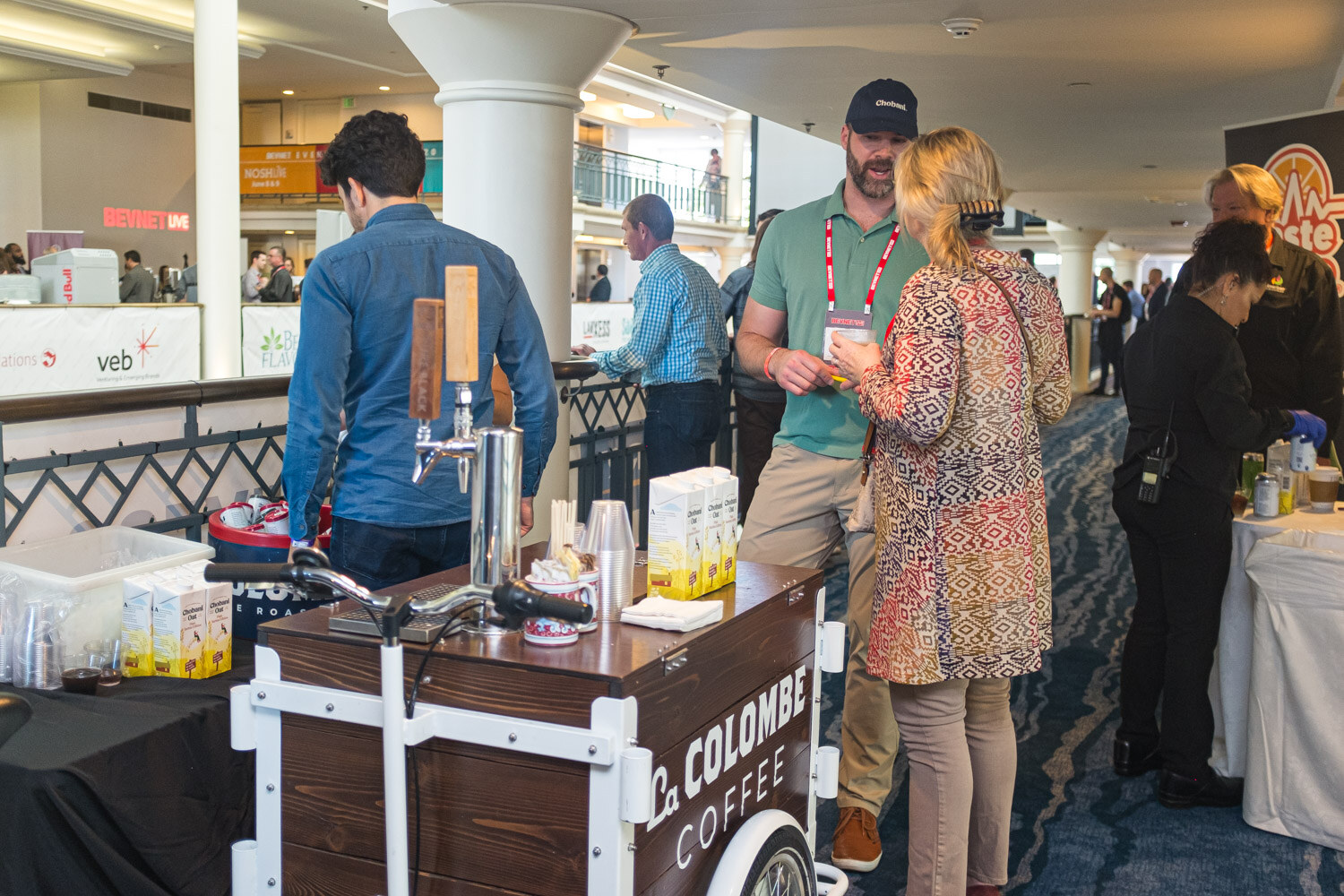 A wooden La Colombe cart with a tap handle stands in the atrium with attendees in conversation around it.