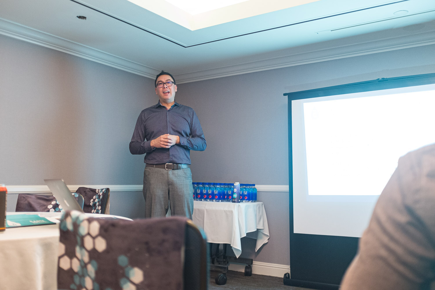 A man in a blue shirt stands beside a projection screen and a table with water bottles while presenting.
