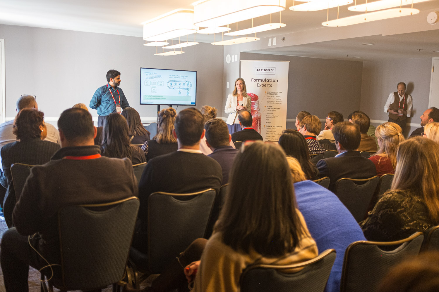 Rows of seated attendees face two presenters standing near a screen and a pull-up banner.
