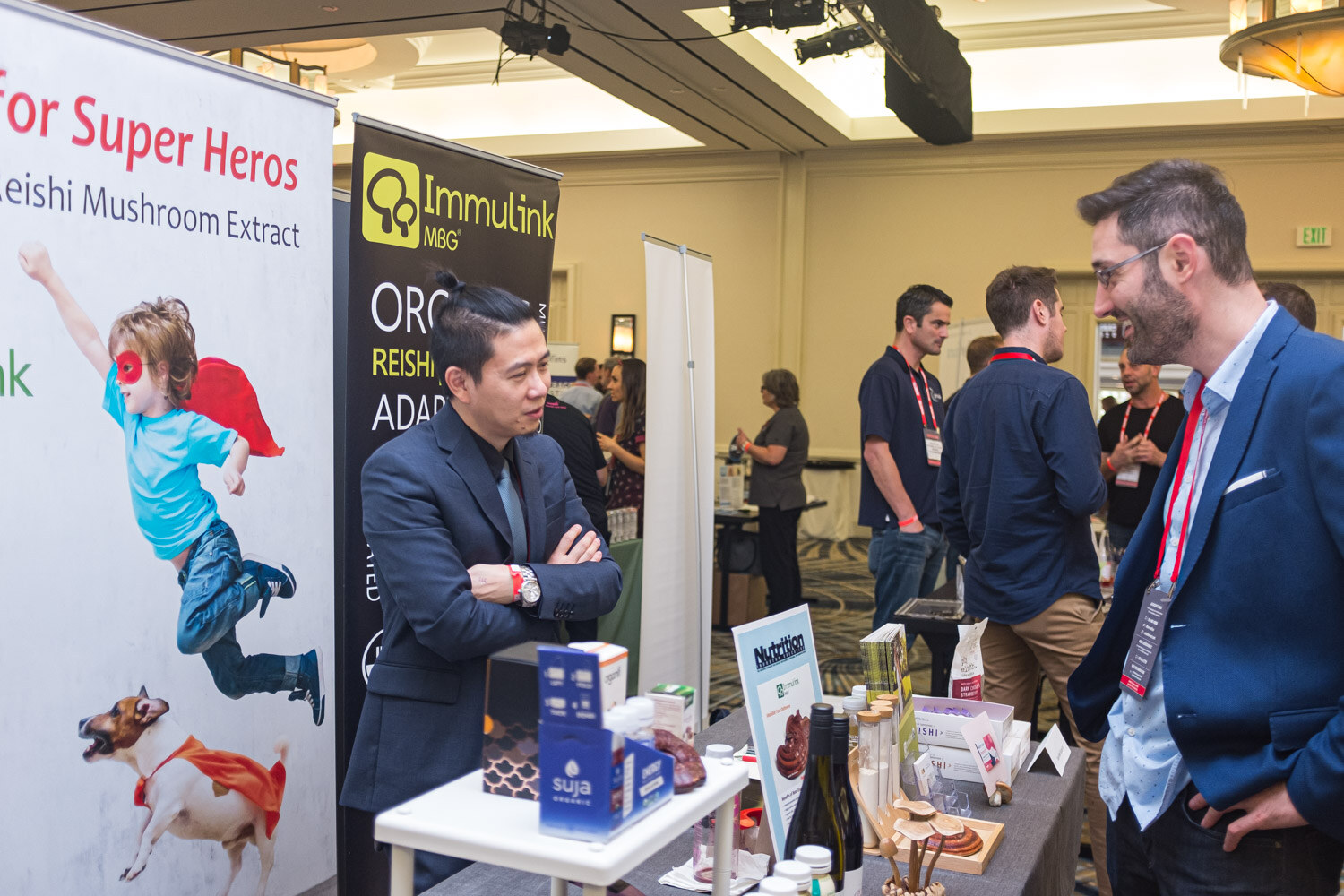 A man in a suit speaks with a man in a blue blazer beside a banner for mushroom extract products.