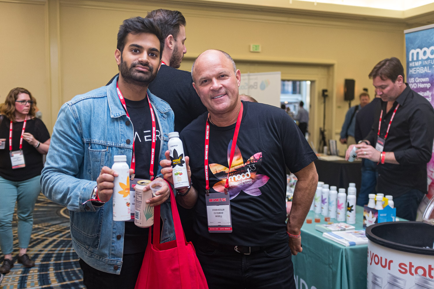 A man in a denim jacket and a man in a black shirt hold bottles and smile in the expo room.